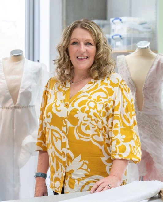 Woman in a yellow and white patterned shirt standing in a fashion studio at Glasgow Kelvin College, with mannequins and fabric on the table. Woman in a yellow and white patterned shirt standing in a fashion studio at Glasgow Kelvin College, with mannequins and fabric on the table.